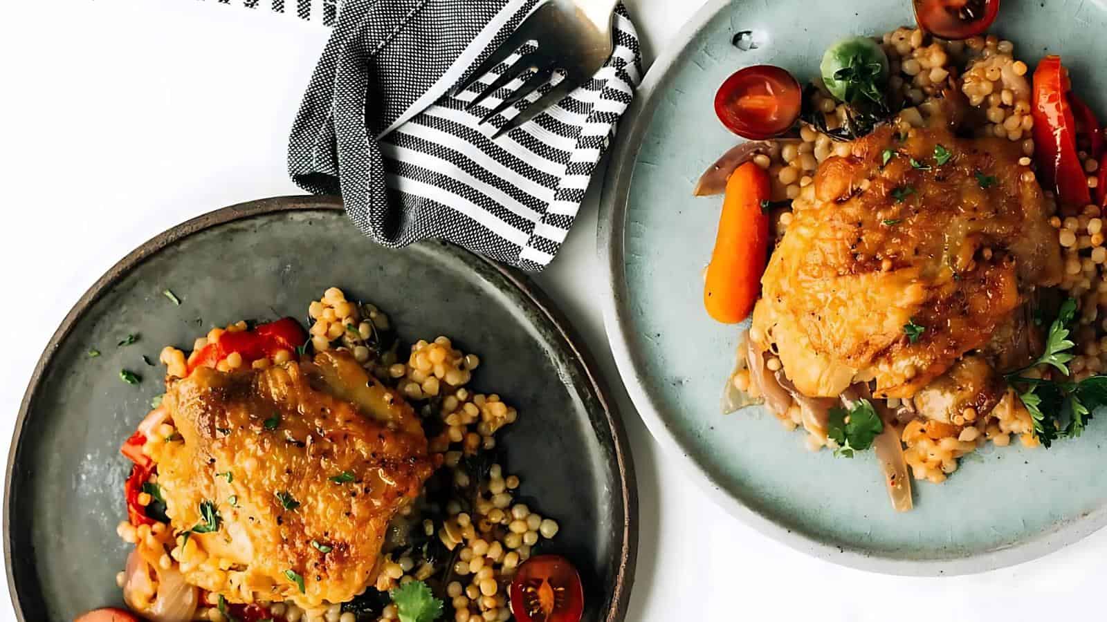 Two plates with couscous, roasted chicken thighs, cherry tomatoes, and carrots, garnished with herbs; a striped napkin and fork are in the background.