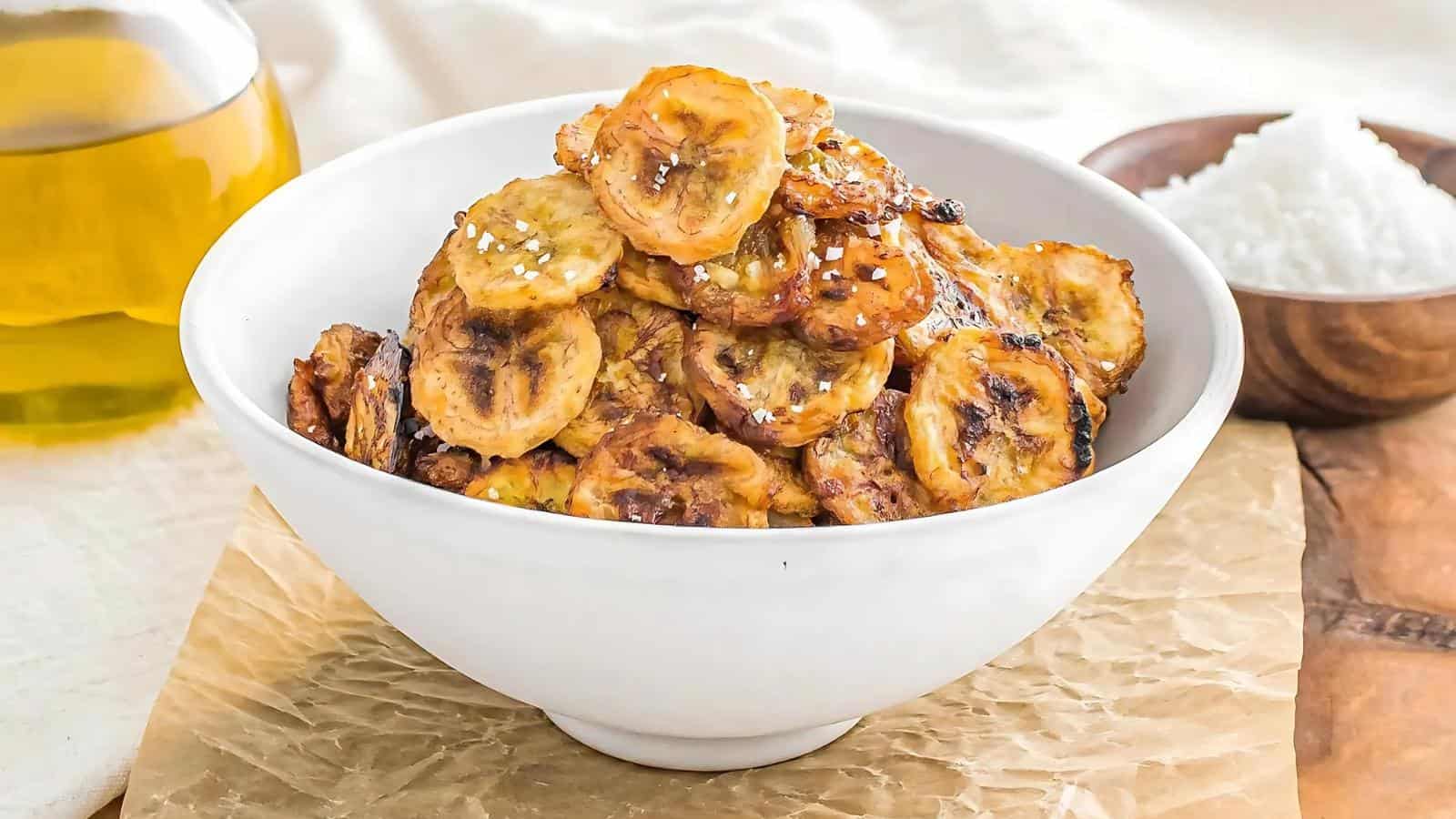 A white bowl filled with grilled or roasted sliced plantains, garnished with sesame seeds. A bottle of oil and a bowl of white rice are visible in the background.