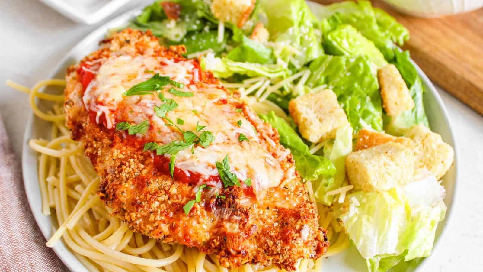 A plate of breaded chicken parmesan on spaghetti, topped with tomato sauce and melted cheese, served with a green salad and croutons.