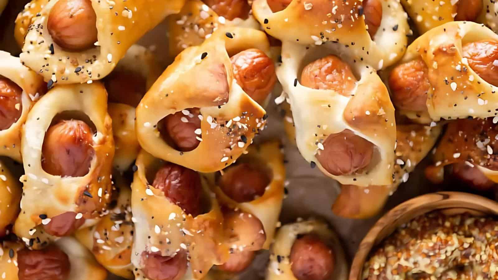Mini sausages wrapped in golden-brown pretzel dough, sprinkled with everything bagel seasoning, next to a bowl of whole grain mustard.