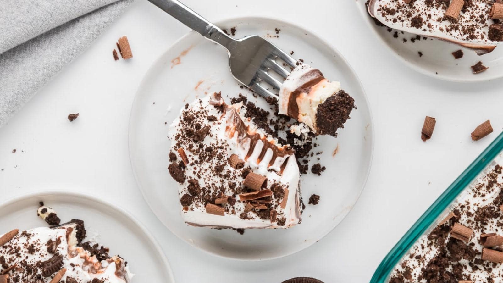 A plate with a slice of layered chocolate dessert topped with whipped cream, chocolate shavings, and crumbs; a fork holds a bite-sized piece.
