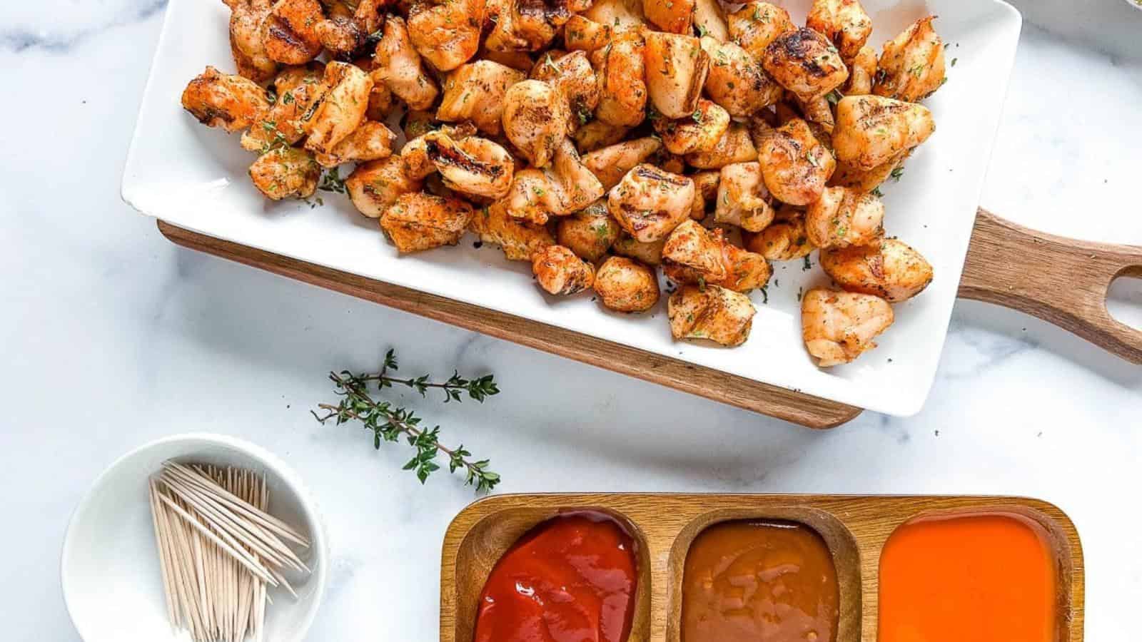 A rectangular plate of grilled, seasoned chicken bites on a wooden board, with three dipping sauces and a bowl of toothpicks beside it.