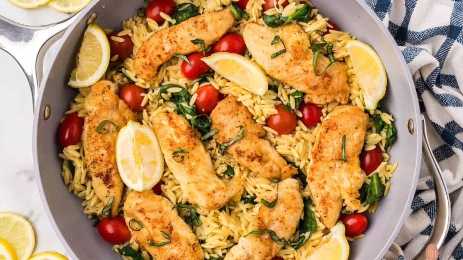A skillet with cooked chicken tenders, orzo pasta, cherry tomatoes, spinach, and lemon wedges, garnished with fresh basil.