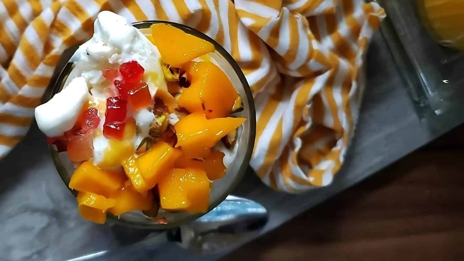 A glass filled with diced yellow fruit, whipped cream, and red jelly pieces, placed on a gray surface with a yellow and white striped cloth nearby.