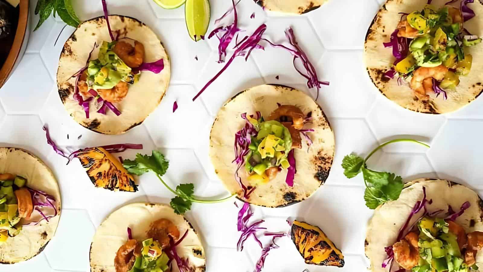 Several tacos on tortillas filled with shrimp, purple cabbage, and green salsa are arranged on a white surface, garnished with cilantro, lime wedges, and grilled pineapple.