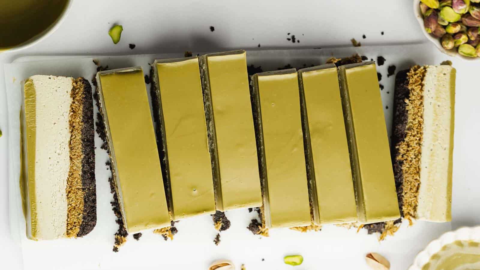 Rectangular slices of a green dessert, likely pistachio-flavored, are arranged on a white tray with scattered pistachios around.