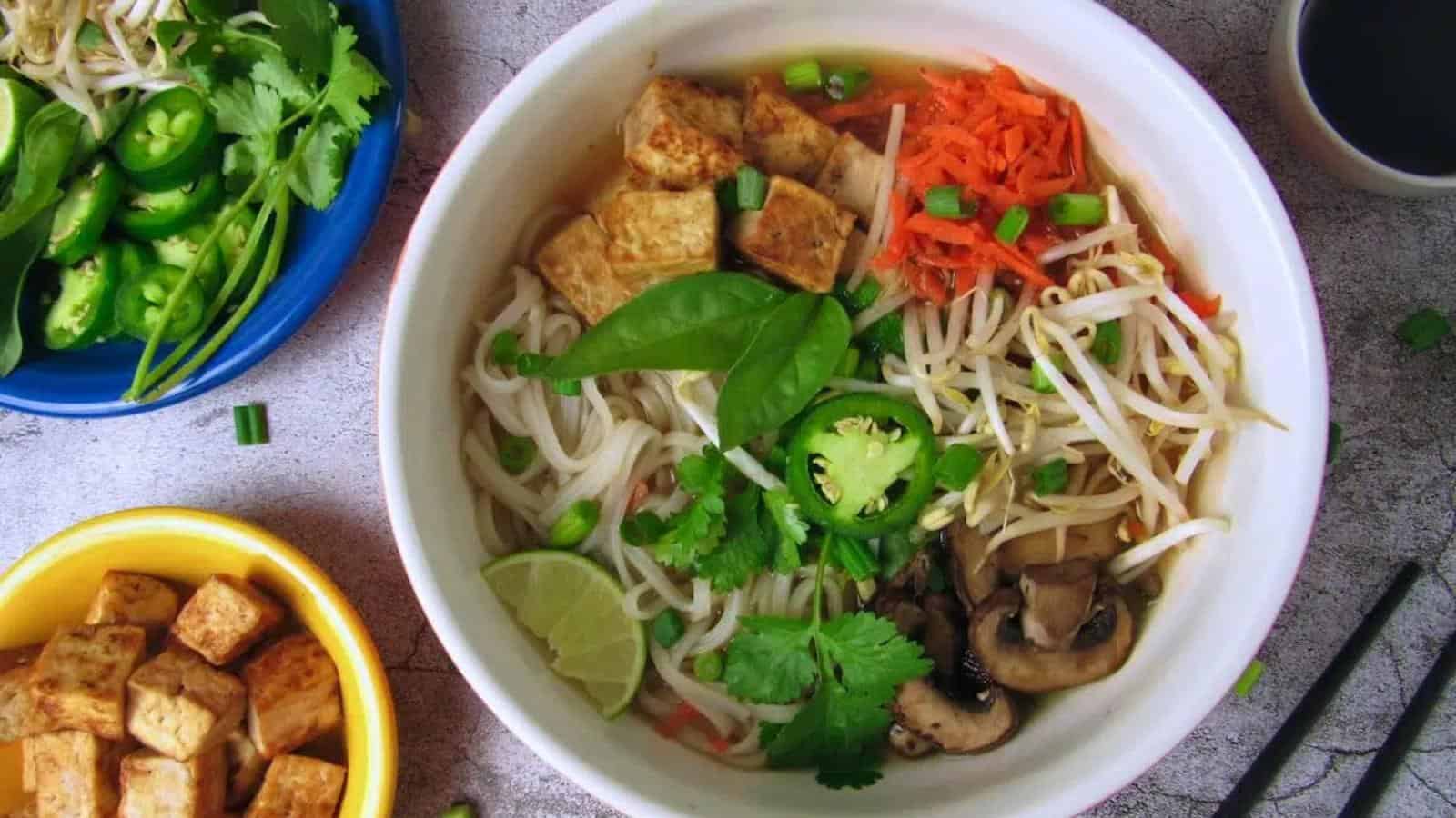 A bowl of noodle soup topped with tofu, bean sprouts, sliced jalapeños, mushrooms, shredded carrots, cilantro, basil, green onions, and a lime wedge, with side dishes nearby.