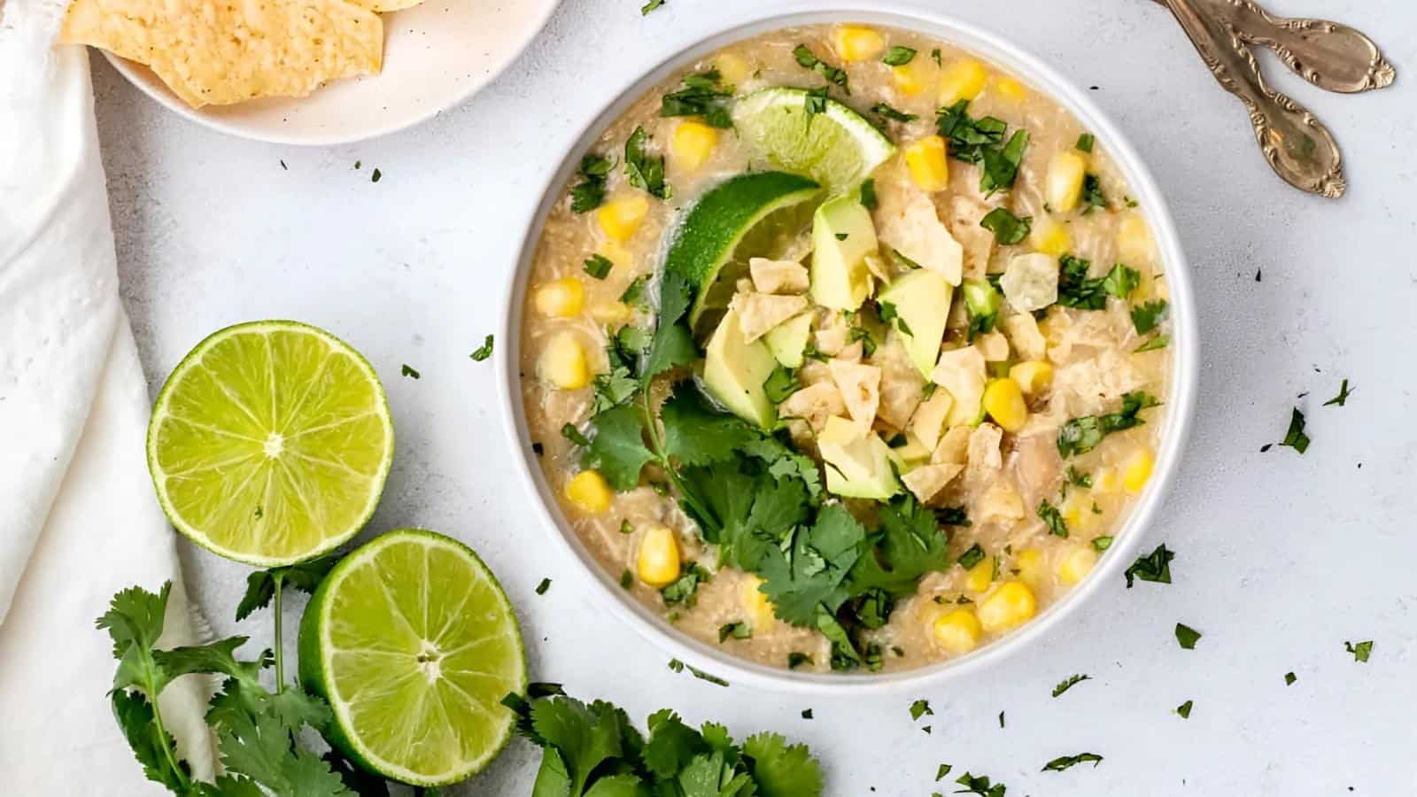 A bowl of chicken soup with corn, avocado, tortilla chips, and cilantro, garnished with lime wedges, sits on a light surface with extra lime and chips on the side.