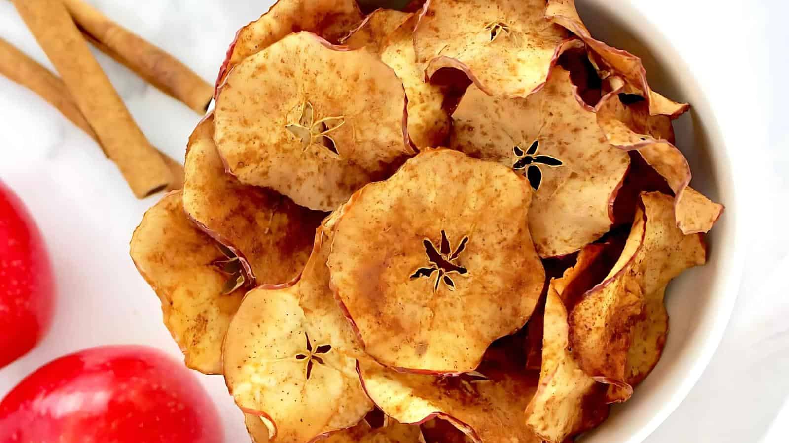 A white bowl filled with dried apple chips, next to whole red apples and cinnamon sticks on a light surface.