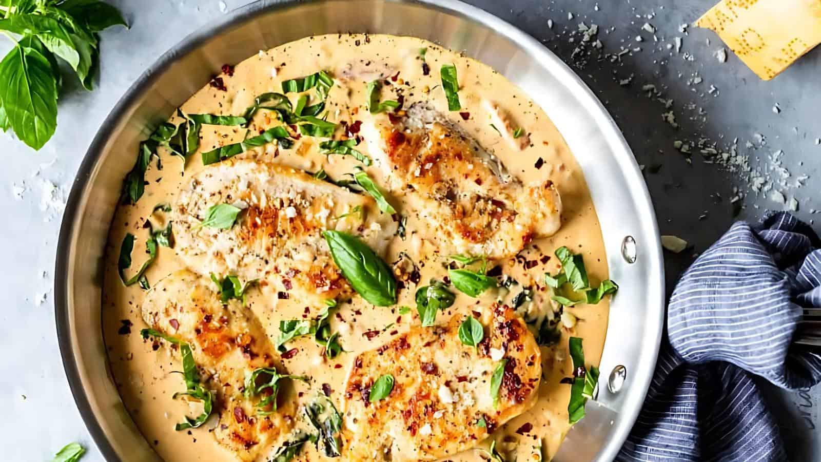 Four pieces of cooked chicken breast in a creamy sauce with herbs and chopped greens in a stainless steel pan, placed on a gray surface.