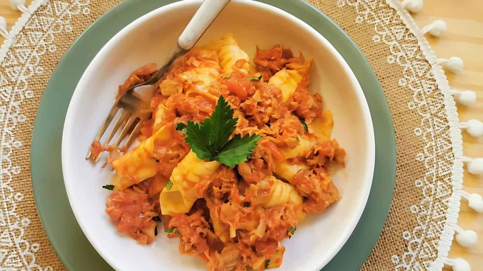 A white bowl filled with pasta topped with a tomato-based sauce and garnished with parsley, placed on a lace-trimmed mat with a fork on the side.