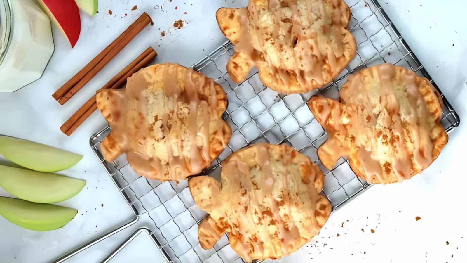 Four leaf-shaped pastries with glaze rest on a cooling rack. Nearby are apple slices, cinnamon sticks, a glass of milk, and crumbs on a white surface.