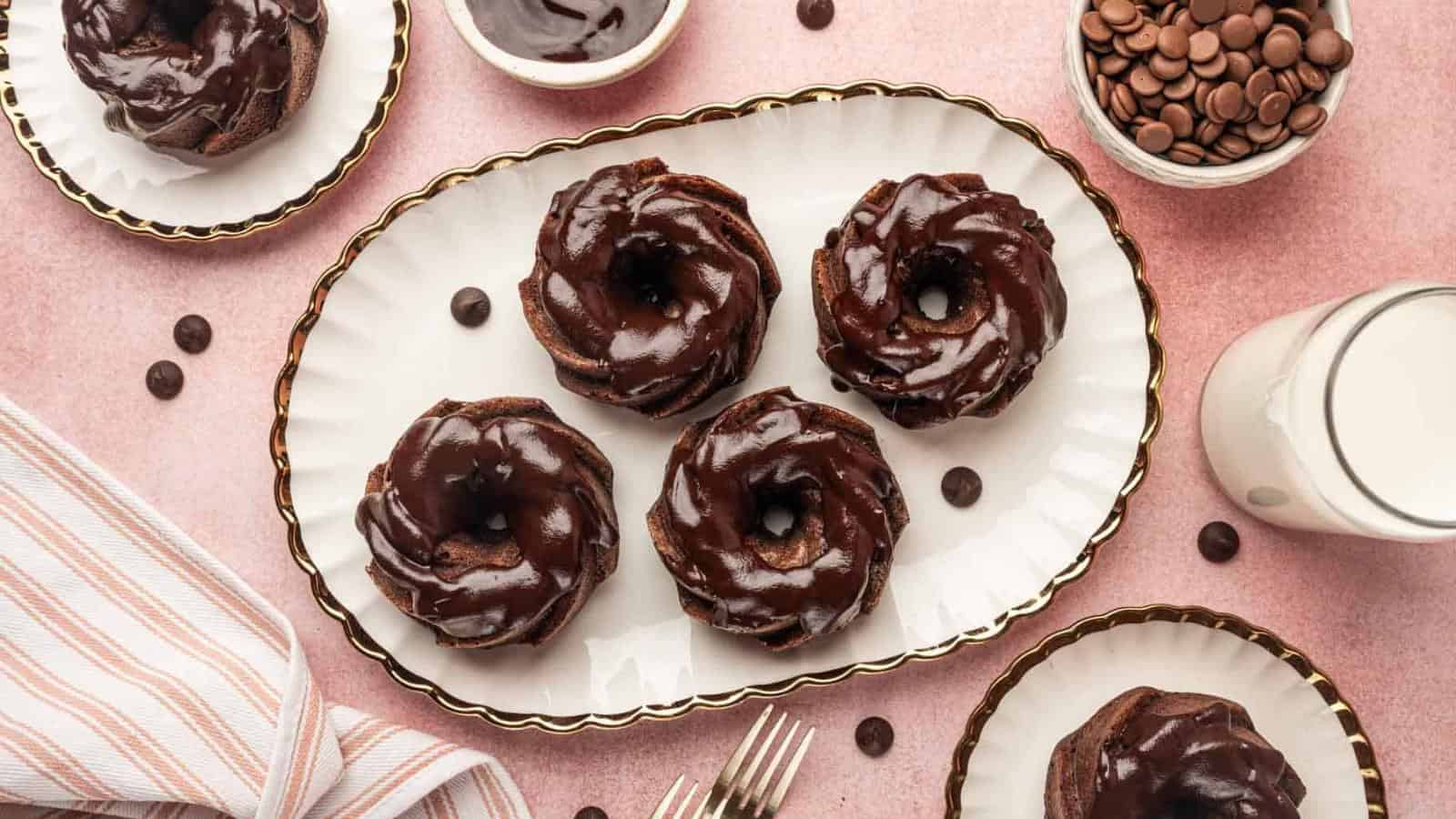 Five chocolate-frosted mini bundt cakes on a white platter, surrounded by bowls of chocolate, a glass of milk, and utensils on a pink surface.