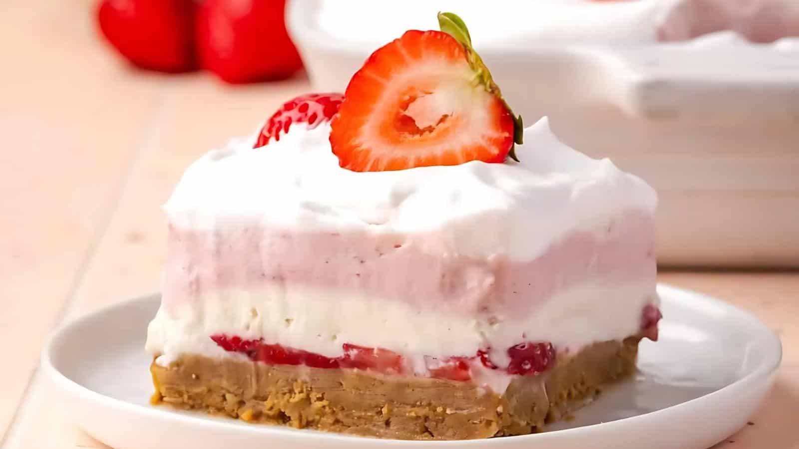 A slice of layered strawberry dessert with a cookie crust, whipped cream, and a sliced strawberry on top, served on a white plate.