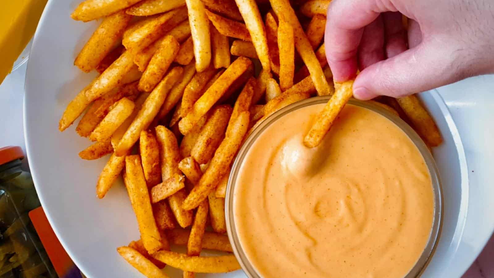 A hand dips a seasoned French fry into a bowl of orange dipping sauce, with more fries arranged on a white plate.