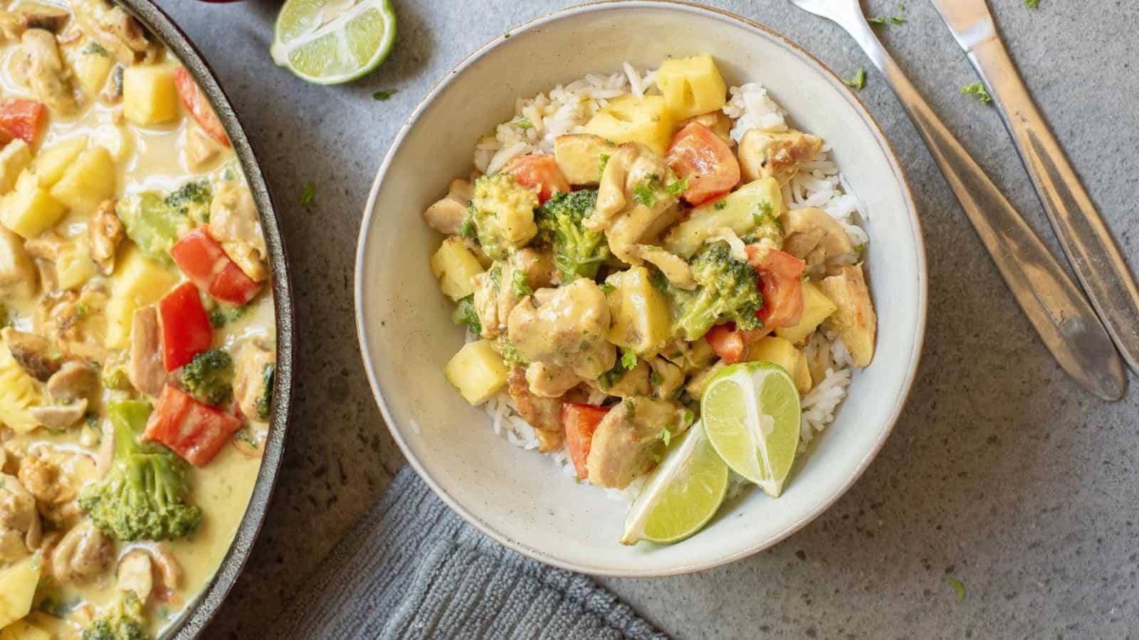 A bowl of rice topped with chicken curry, broccoli, red bell peppers, pineapple, and lime wedges sits next to a skillet with more curry and a set of utensils.