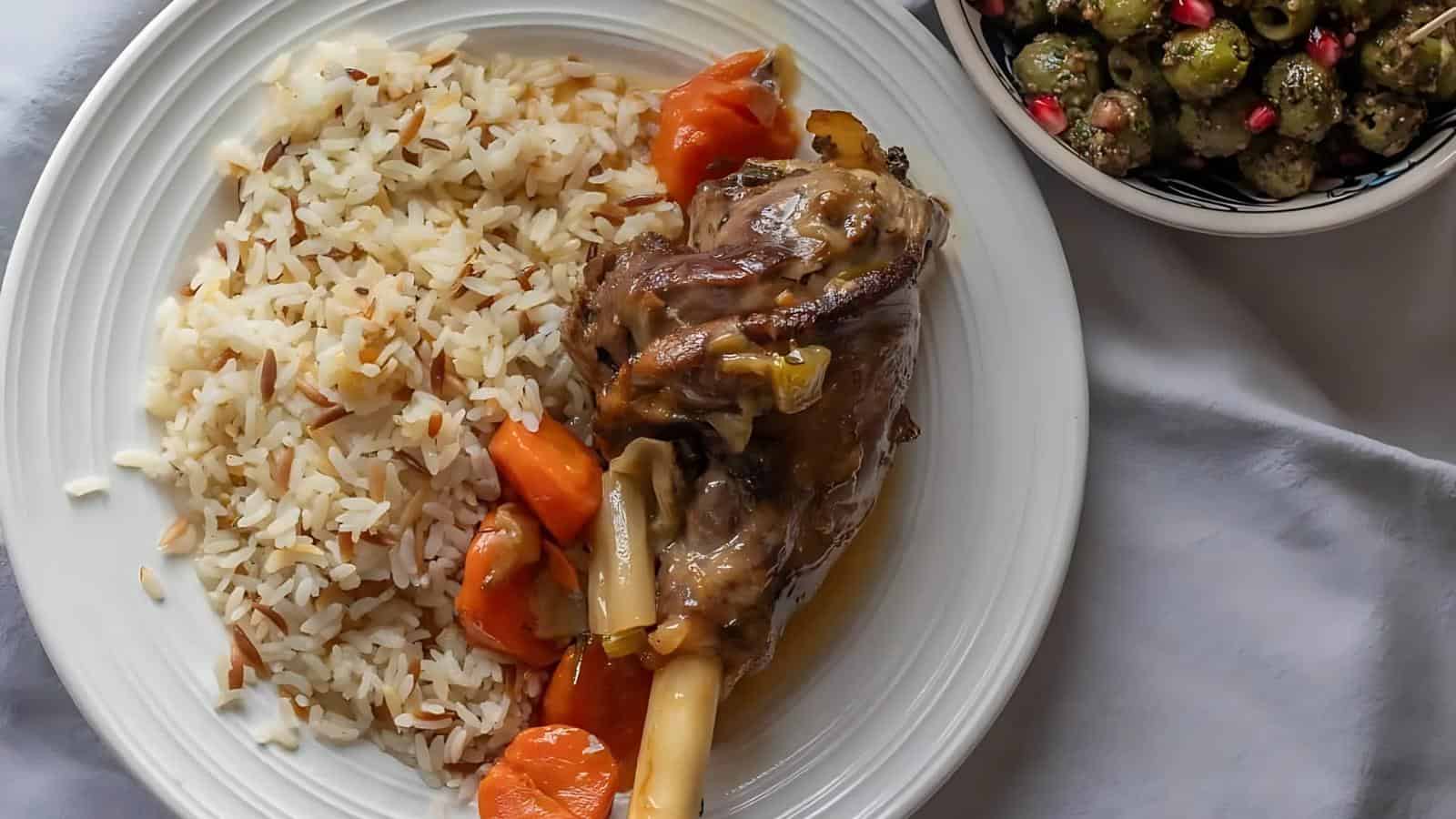 A plate with rice, roasted lamb shank, and carrots, next to a bowl of cooked Brussels sprouts garnished with pomegranate seeds.
