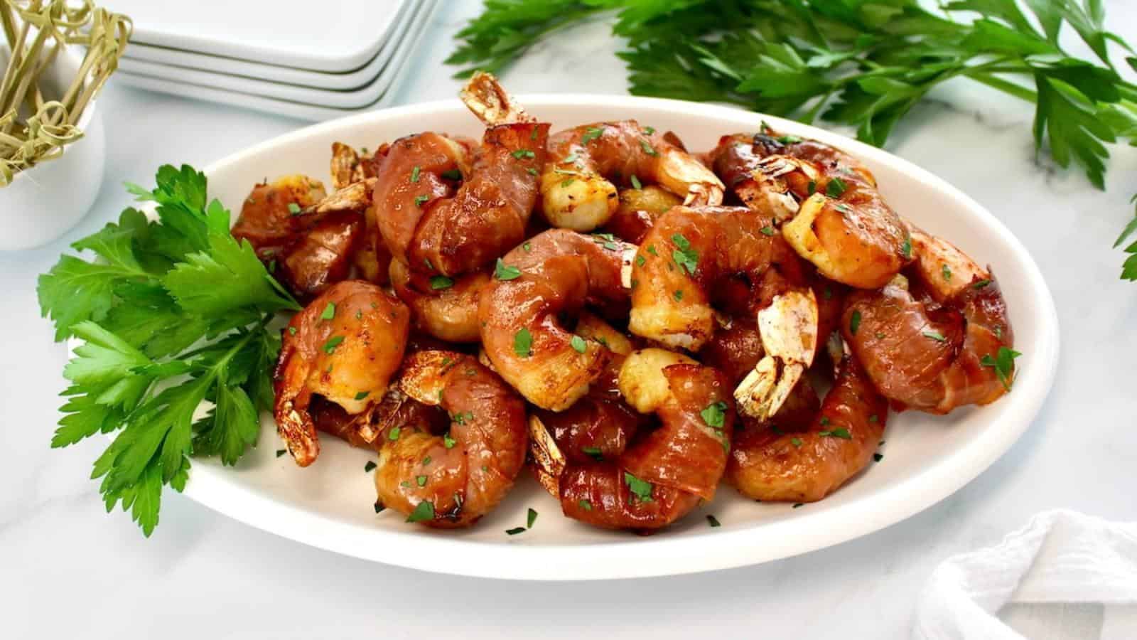 A white oval plate filled with bacon-wrapped shrimp, garnished with chopped herbs and fresh parsley, sits on a white surface.
