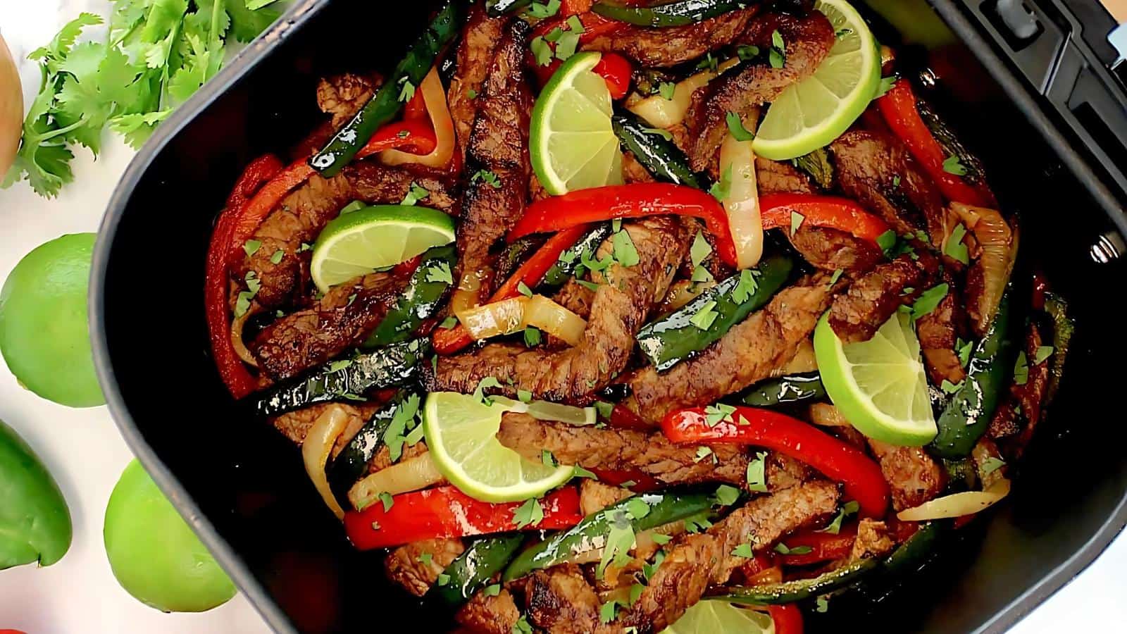 Sliced beef, red and green bell peppers, onions, and lime wedges cooked together in an air fryer basket, garnished with chopped cilantro.
