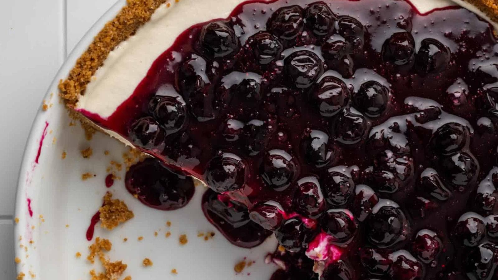 A close-up of a blueberry cheesecake with a graham cracker crust, showing a missing slice on a white plate.