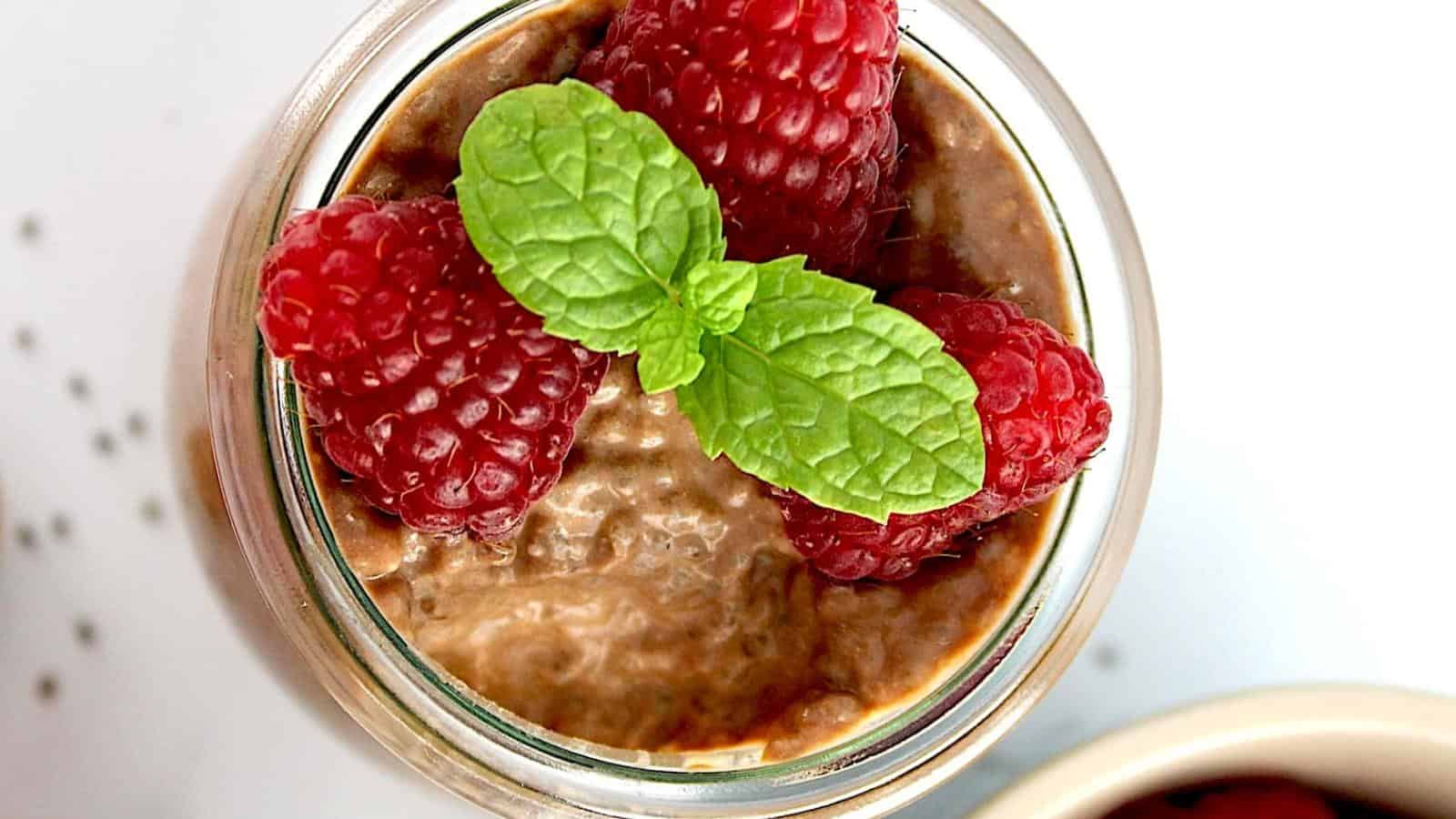 A glass jar filled with chocolate chia pudding, topped with three fresh raspberries and a sprig of mint.