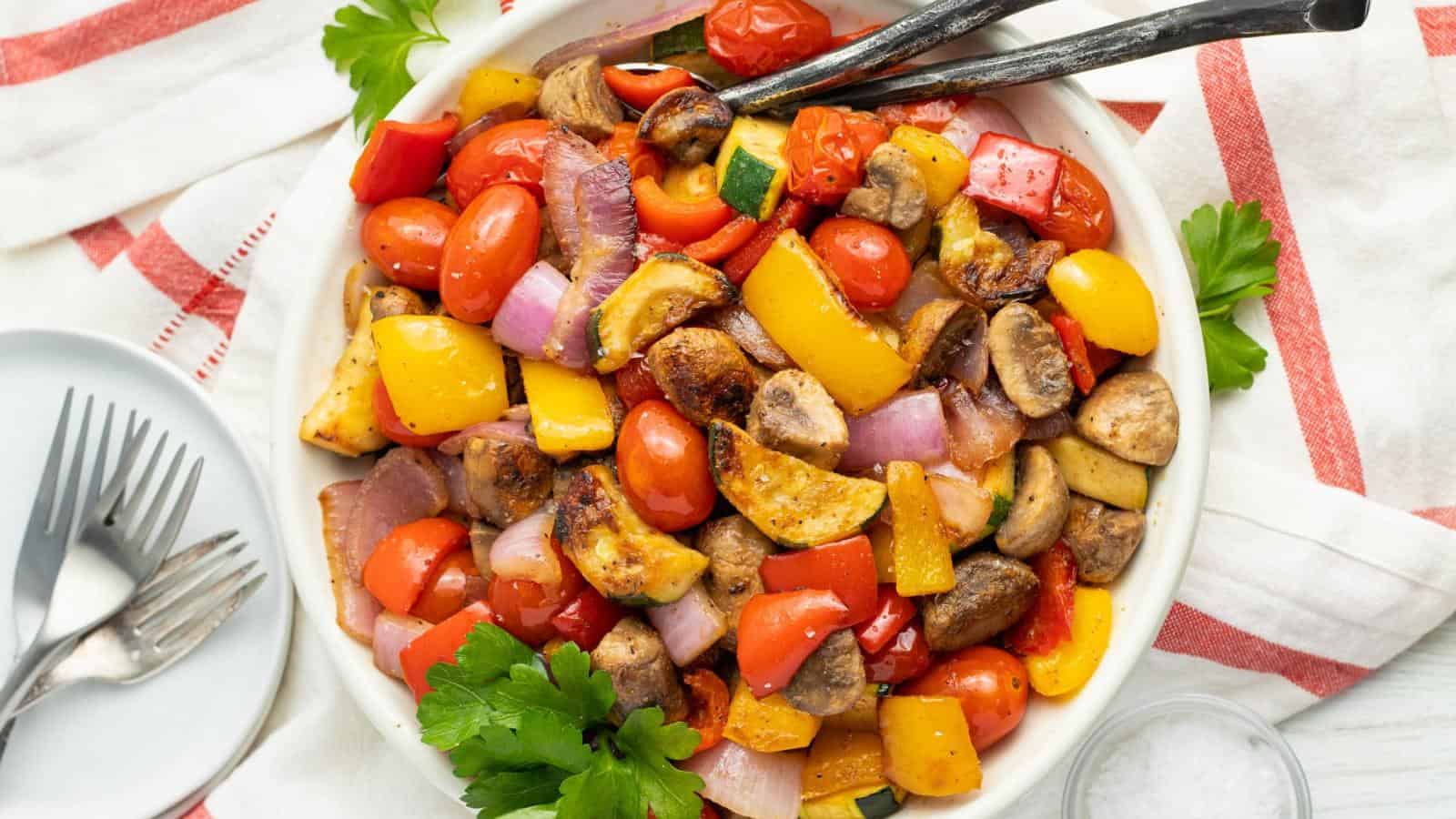 A bowl of roasted vegetables, including cherry tomatoes, mushrooms, red onions, yellow and red bell peppers, garnished with parsley, on a table with a red-striped napkin.