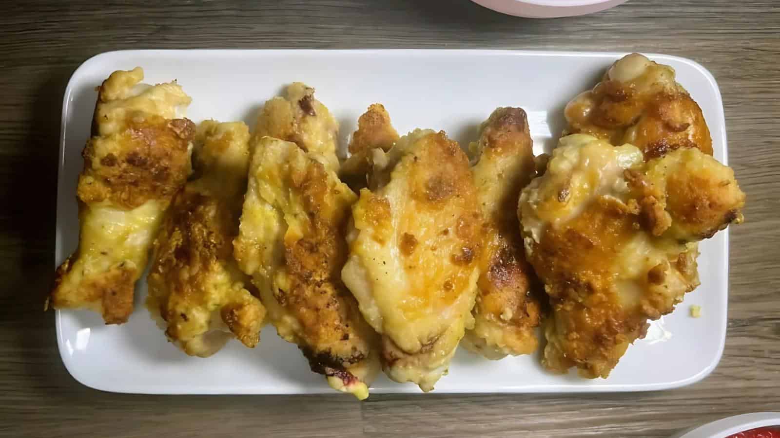 Six pieces of baked or fried chicken wings arranged in a row on a rectangular white plate set on a wooden surface.