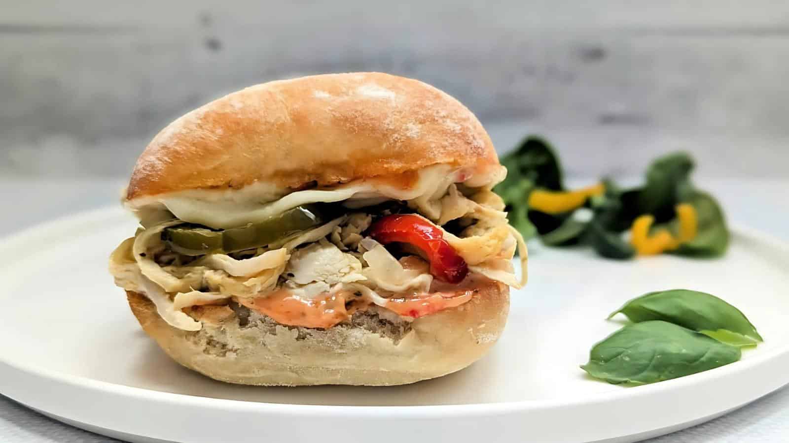 A sandwich with shredded chicken, melted cheese, pickles, and red peppers on a bun, served on a white plate with greens and basil leaves in the background.