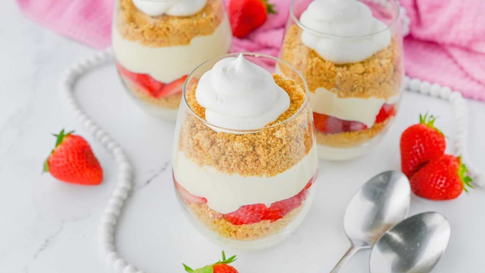 Three dessert glasses layered with whipped cream, crushed cookies, and sliced strawberries, placed on a white tray with fresh strawberries and two spoons beside them.