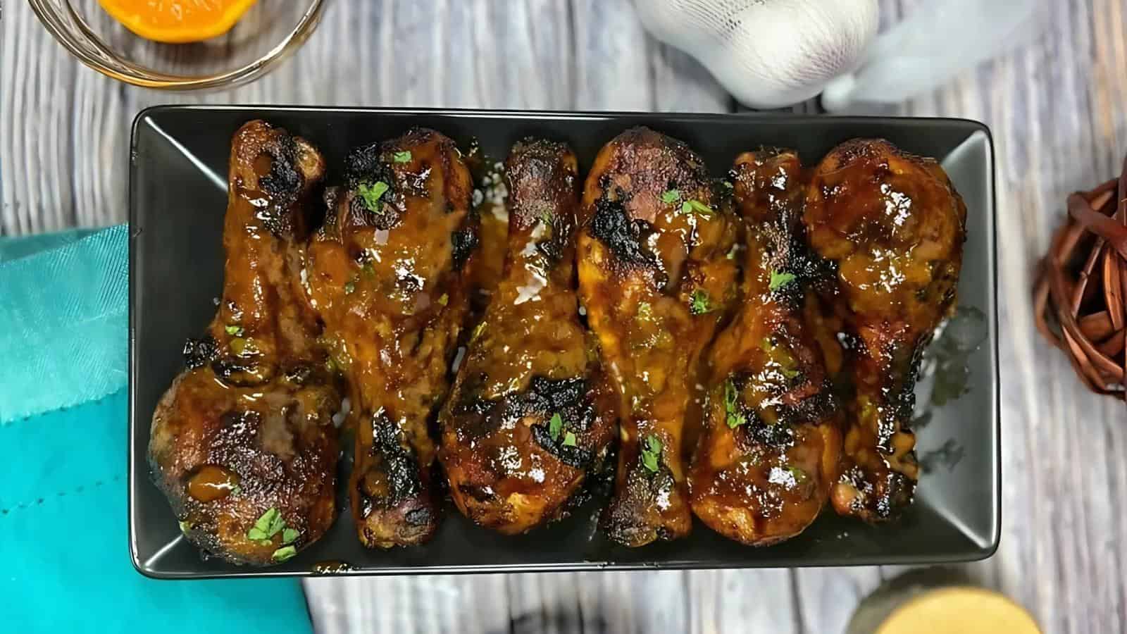 A rectangular black plate with seven grilled chicken drumsticks coated in a brown sauce and garnished with chopped herbs, placed on a wooden surface.