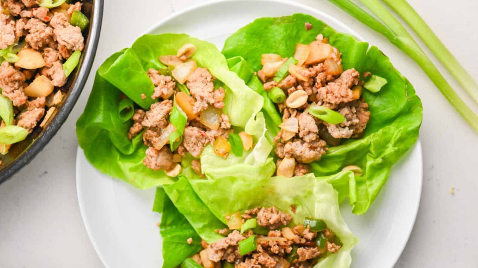 Three lettuce wraps filled with ground meat, chopped vegetables, and peanuts are arranged on a white plate, with green onions and a skillet visible nearby.