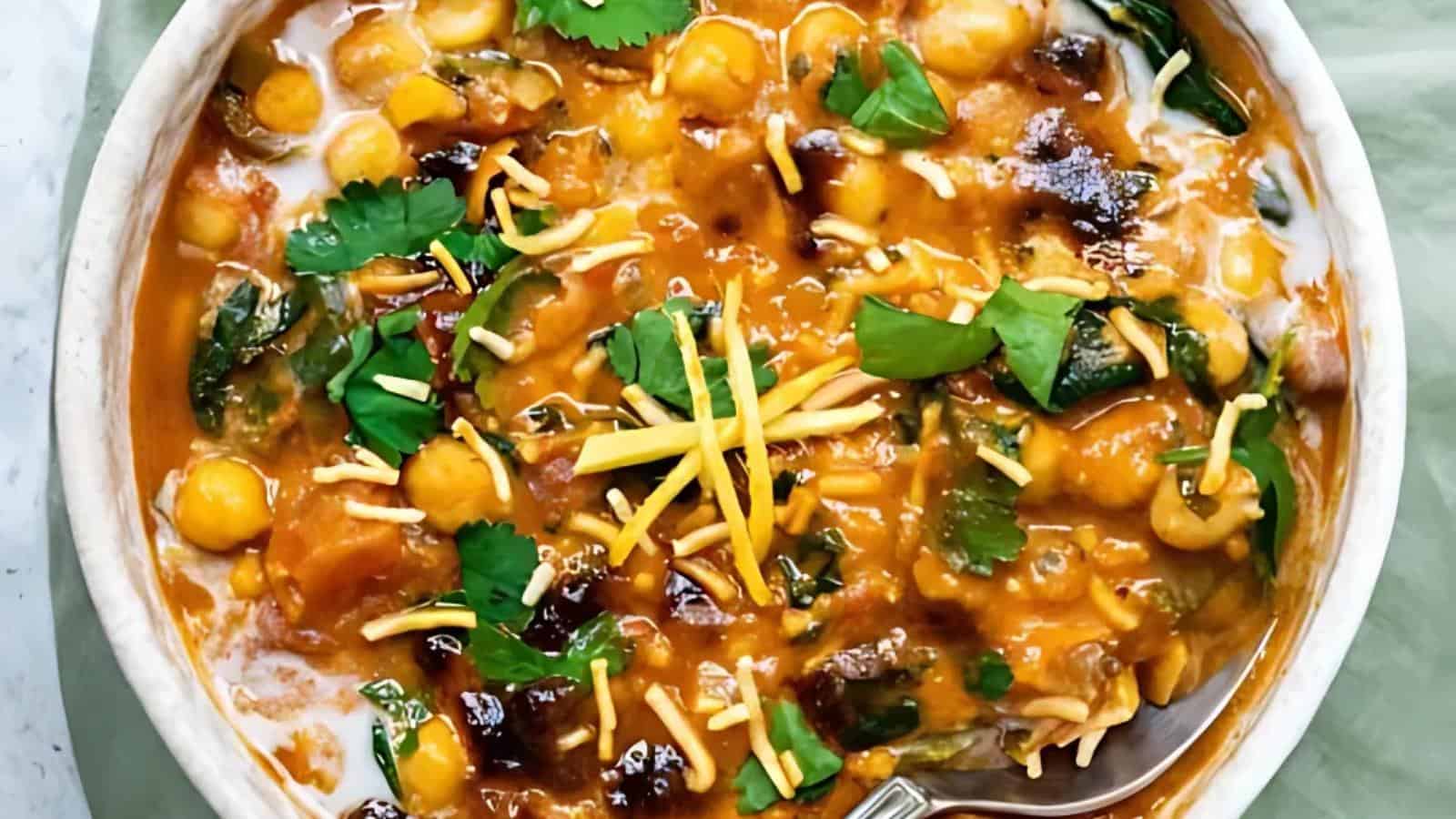 A bowl of chickpea curry topped with shredded cheese, chopped cilantro, and a swirl of cream, with a spoon partially visible.