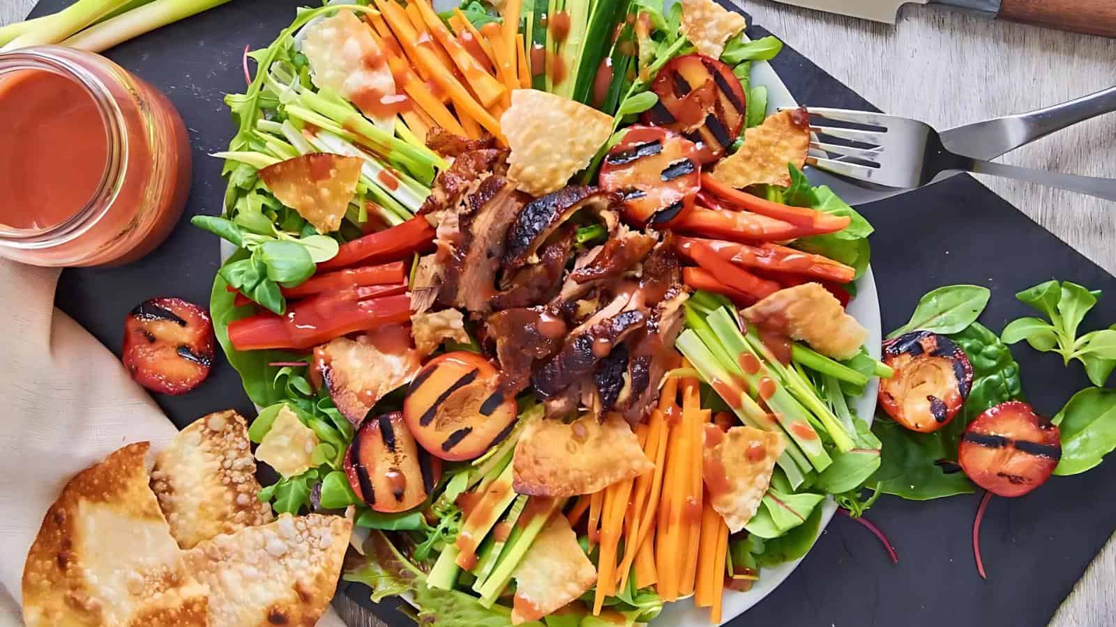 A colorful salad with grilled chicken, sliced vegetables, grilled peaches, tortilla chips, and dark dressing, served on a white plate with a jar of sauce on the side.
