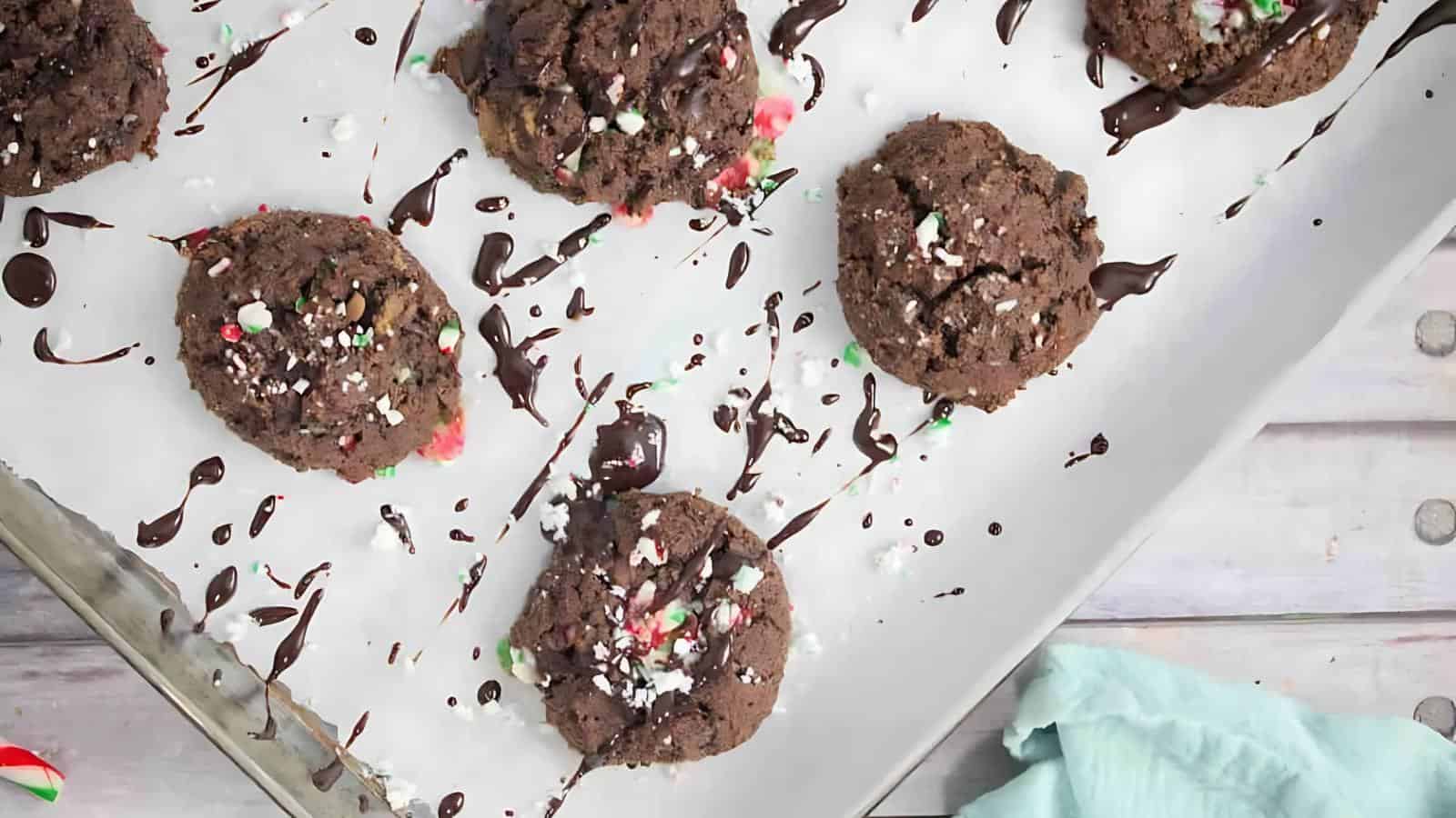 Six chocolate cookies topped with crushed peppermint and chocolate drizzle are arranged on a parchment-lined baking sheet. A blue cloth is partially visible in the lower right corner.