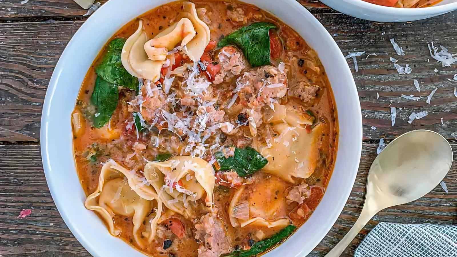 A bowl of creamy soup with tortellini, ground meat, spinach, diced tomatoes, and grated cheese on top, placed on a wooden table next to a spoon.