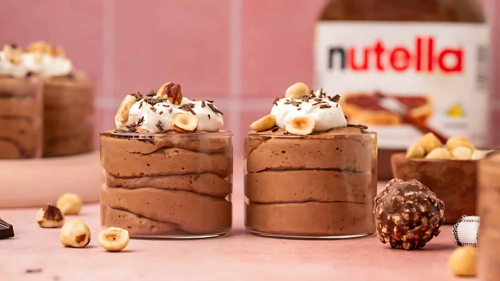 Two glass cups filled with chocolate mousse topped with whipped cream, hazelnuts, and chocolate shavings, with a Nutella jar and hazelnut chocolates in the background.