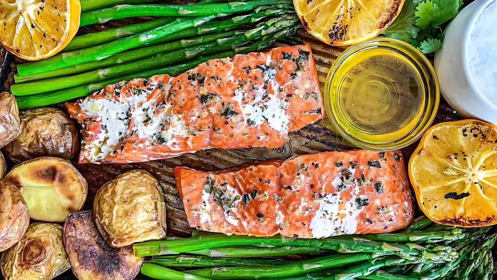 Two cooked salmon fillets with herbs, surrounded by asparagus, roasted potatoes, lemon slices, and a small bowl of oil or sauce on a wooden surface.