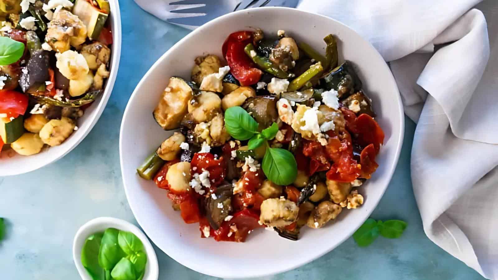 A bowl of mixed salad with chickpeas, tomatoes, eggplant, green beans, crumbled cheese, and fresh basil, placed on a blue surface next to a white napkin.