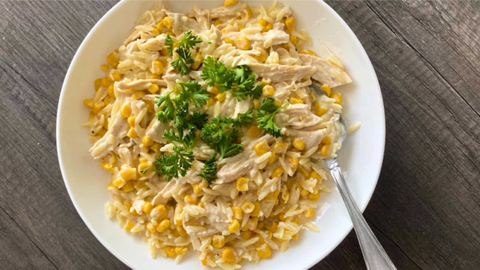 A white bowl filled with creamy pasta, shredded chicken, corn, and parsley garnish sits on a wooden table with a spoon.