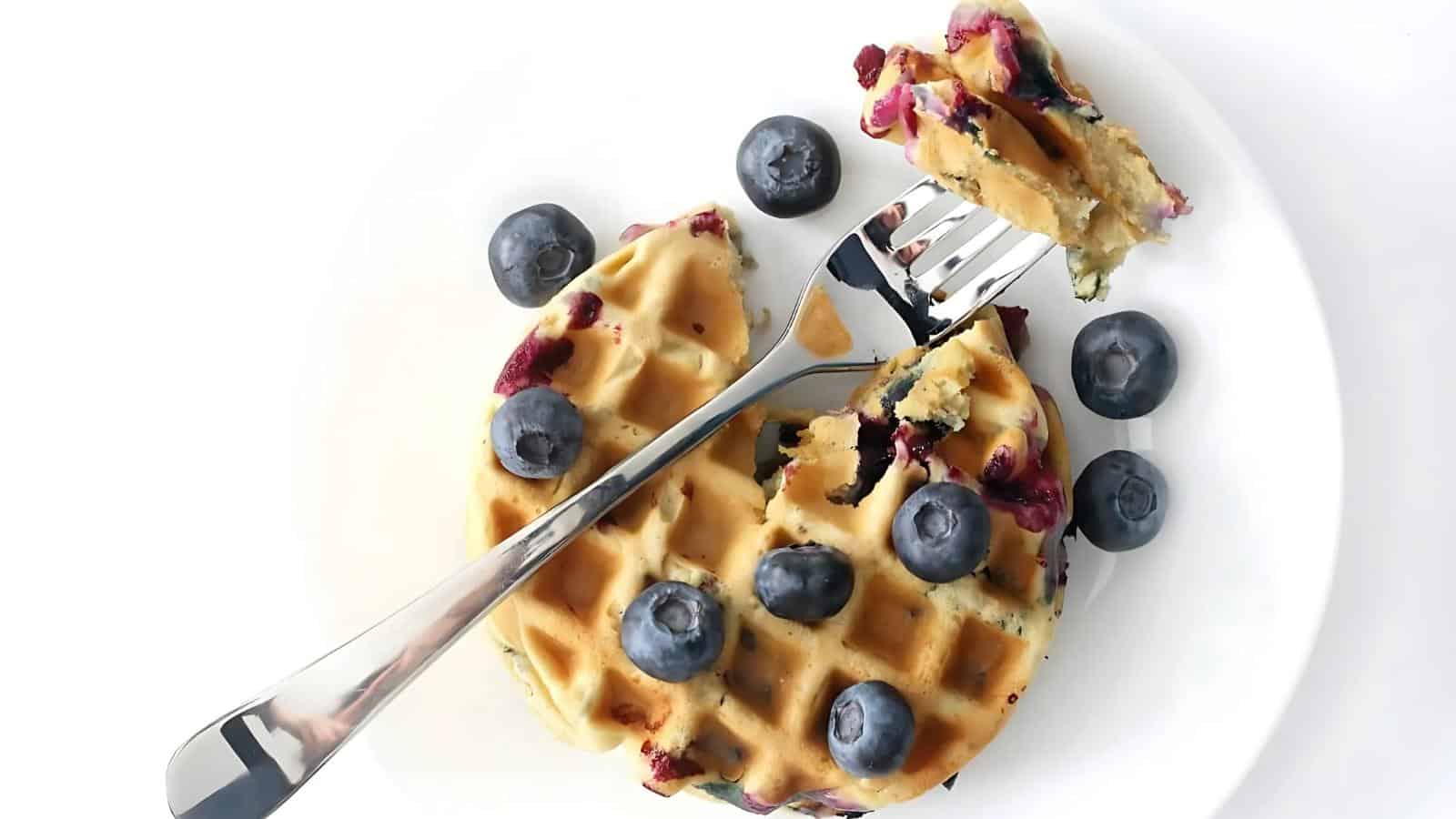 A plate with blueberry waffles topped with fresh blueberries, a fork holding a bite, and a knife resting on the side.