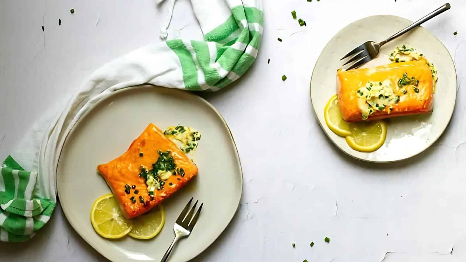 Two plates with baked salmon fillets topped with herbs, garnished with lemon slices; utensils and a green-striped cloth napkin are also visible.