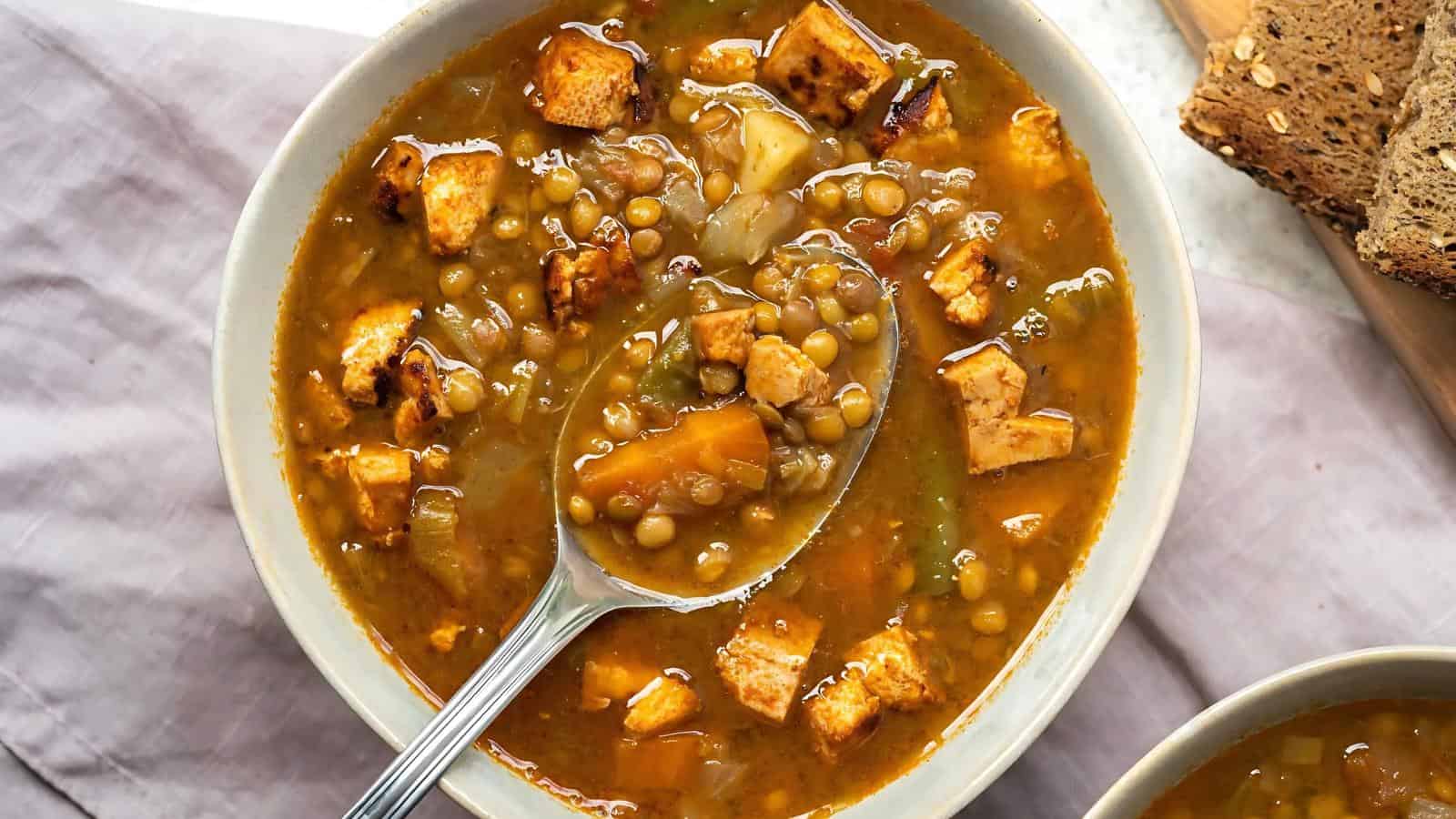 A bowl of lentil soup with diced tofu, carrots, and vegetables, with a spoon lifting some soup; slices of bread are on the side.