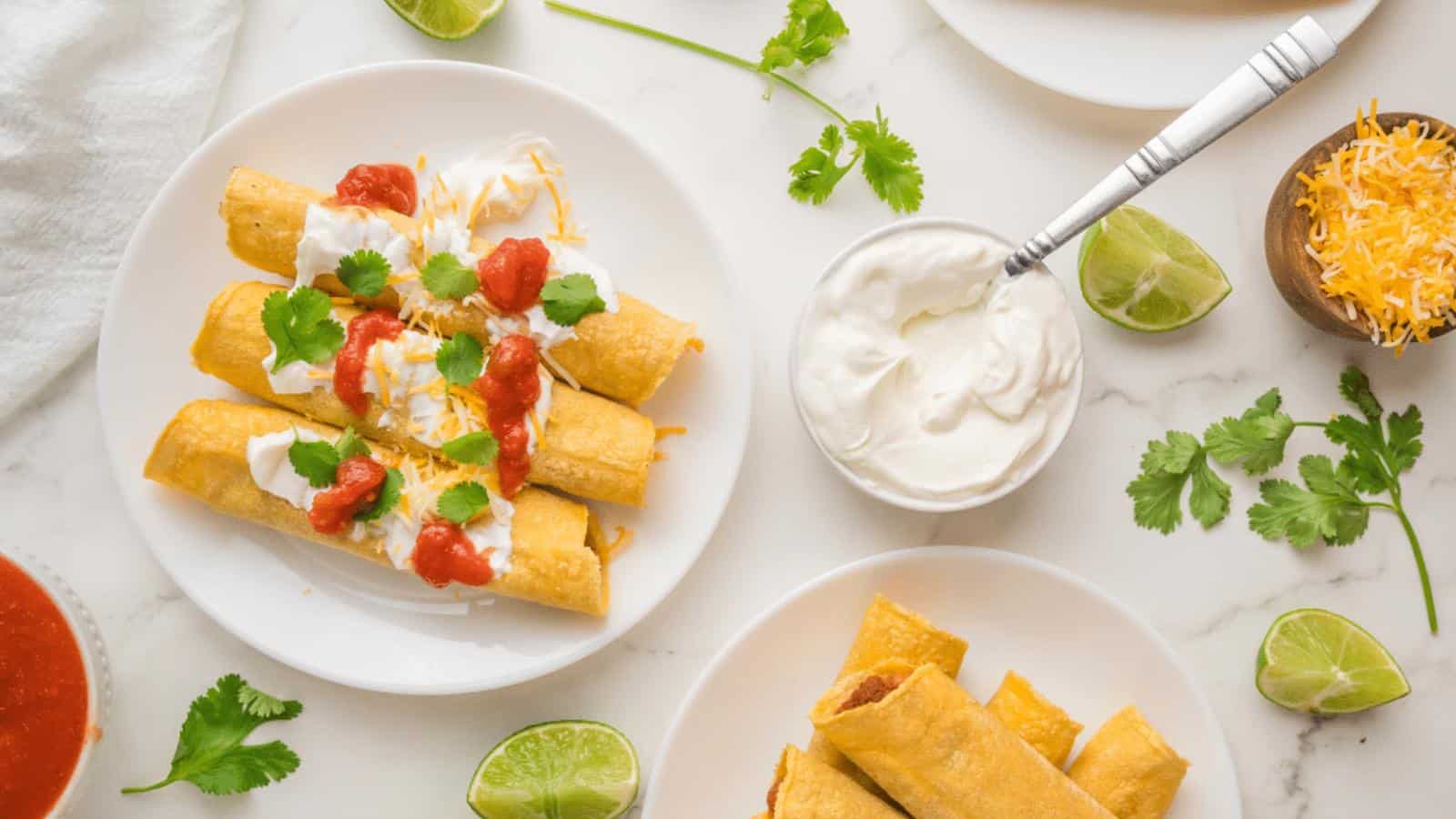 Plates of taquitos topped with sour cream, salsa, shredded cheese, and cilantro are arranged on a table with lime wedges, extra sour cream, cilantro, and a bowl of cheese.