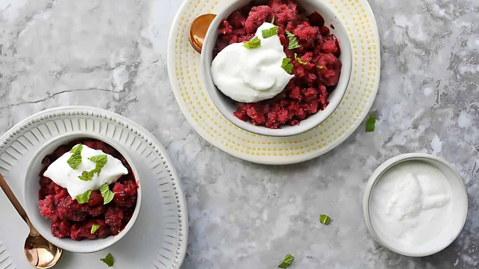 Two bowls of red berry granita topped with whipped cream and mint leaves, served on plates, with an extra bowl of whipped cream on a marble surface.