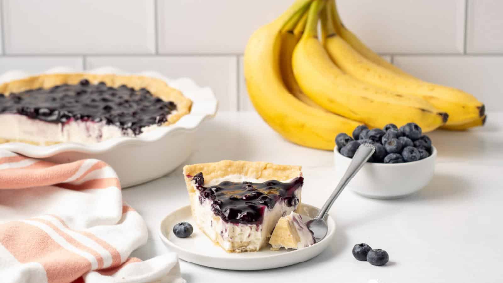 A slice of blueberry-topped pie on a plate with a fork, next to a bowl of blueberries and a bunch of bananas, with the whole pie in the background.