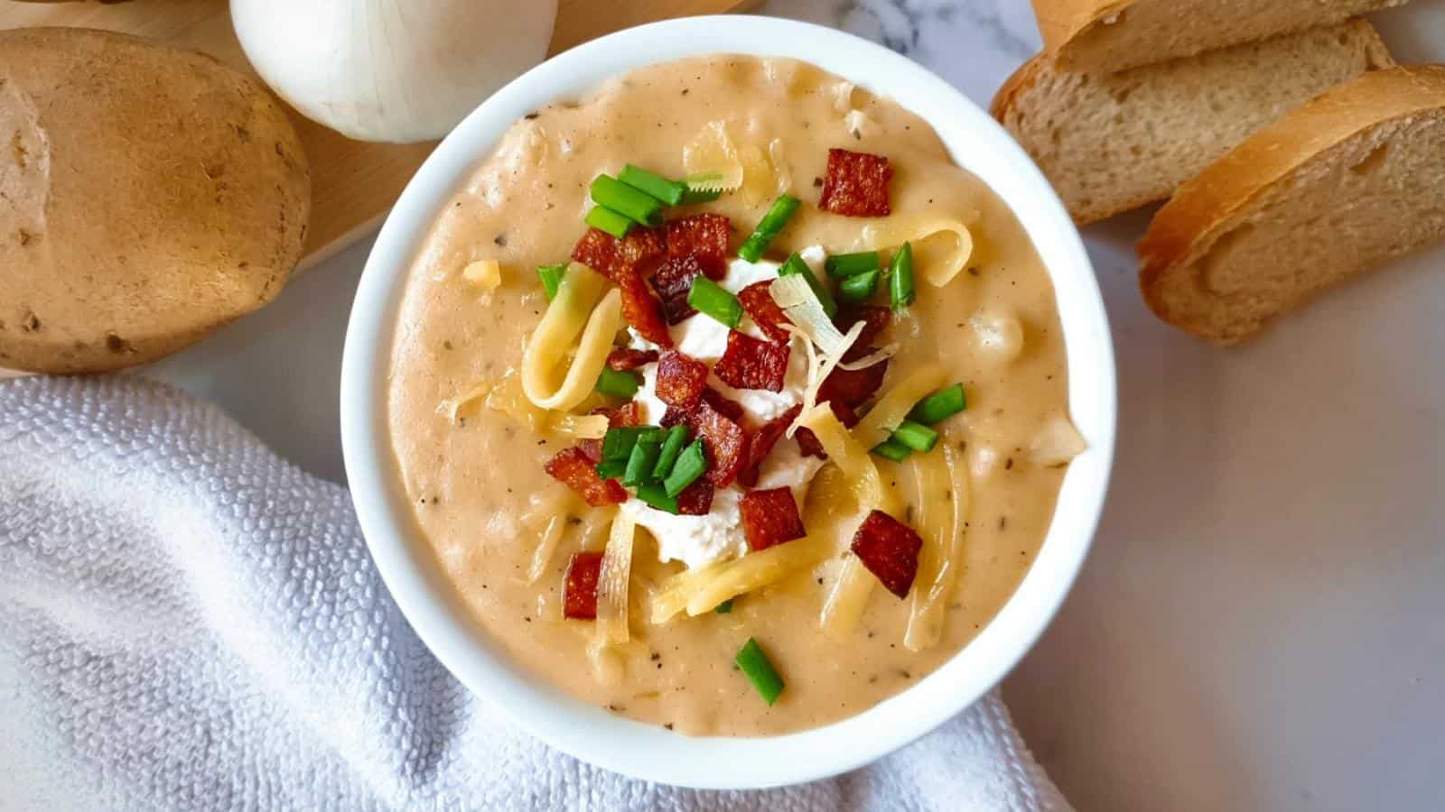 A bowl of creamy soup topped with cheese, bacon bits, and chopped green onions, with bread slices on the side.