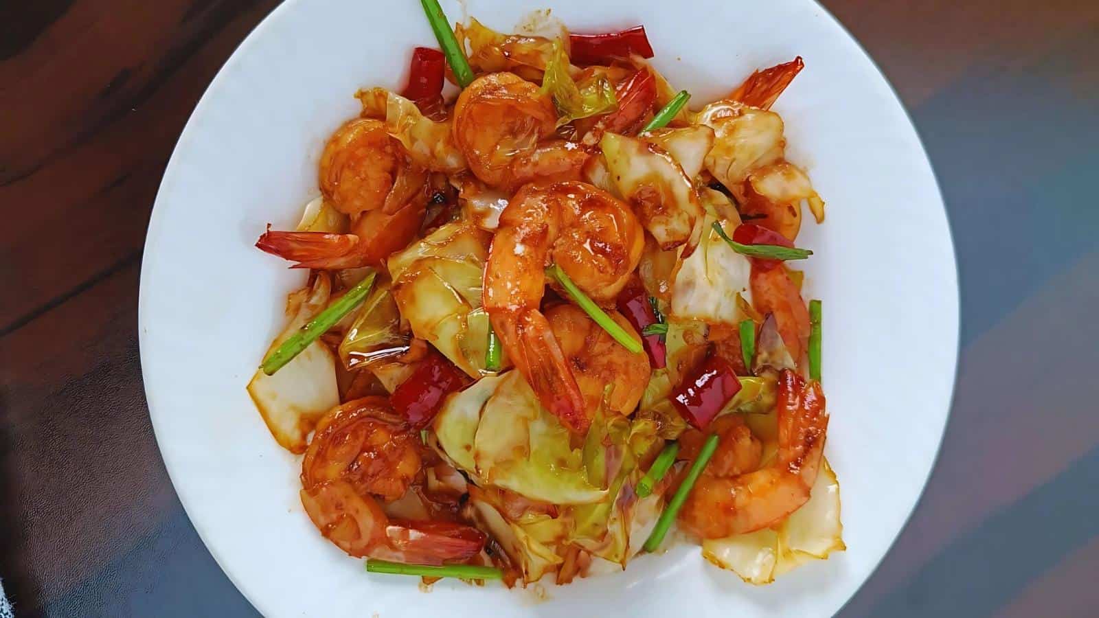 A white plate of stir-fried shrimp with cabbage, red chili peppers, and green onions in a brown sauce.