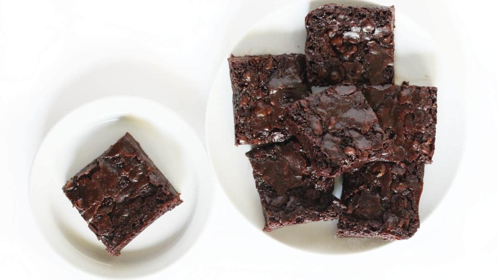 Two white plates on a white surface, one with a single chocolate brownie and the other with a stack of six chocolate brownies.
