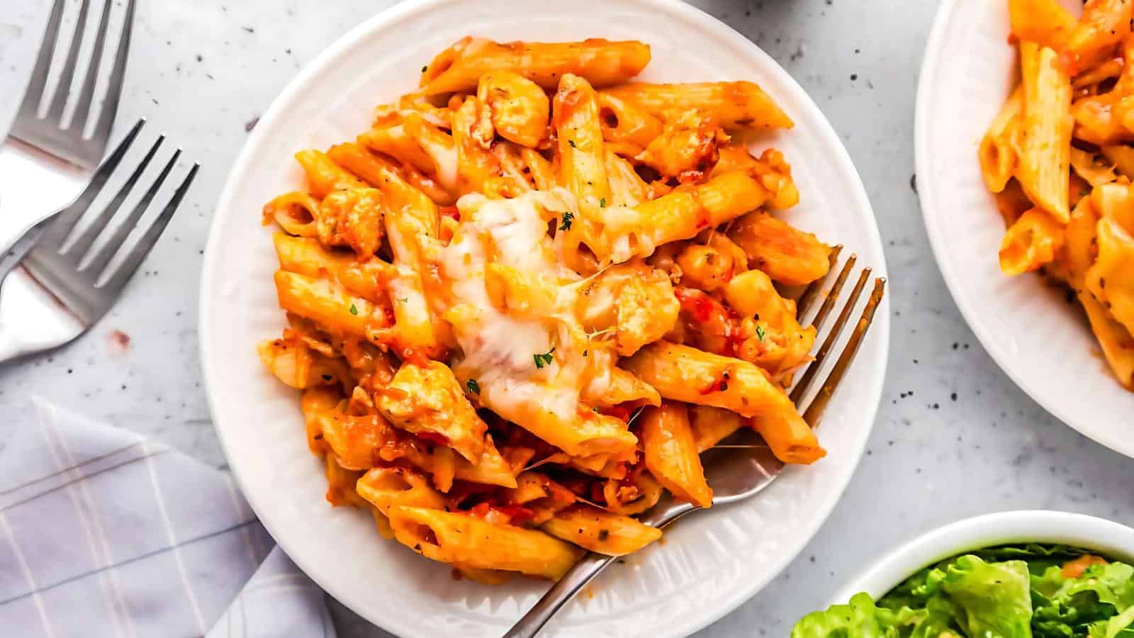 A plate of baked penne pasta with melted cheese and herbs, served with a fork on a white plate.