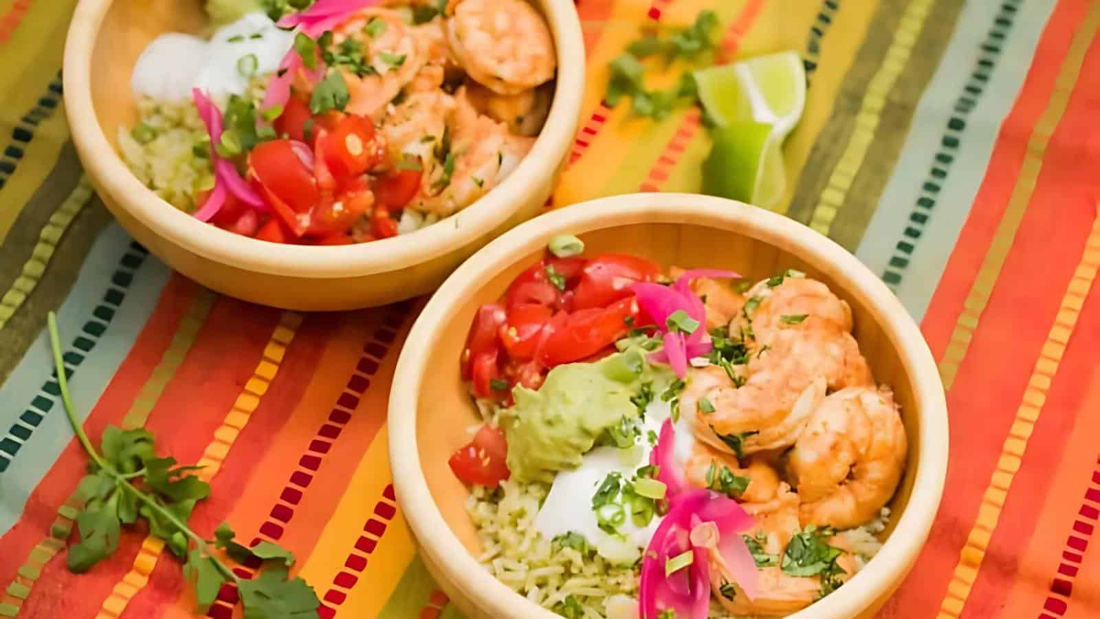 Two wooden bowls filled with shrimp, rice, guacamole, sour cream, tomatoes, and pickled onions on a colorful striped tablecloth.